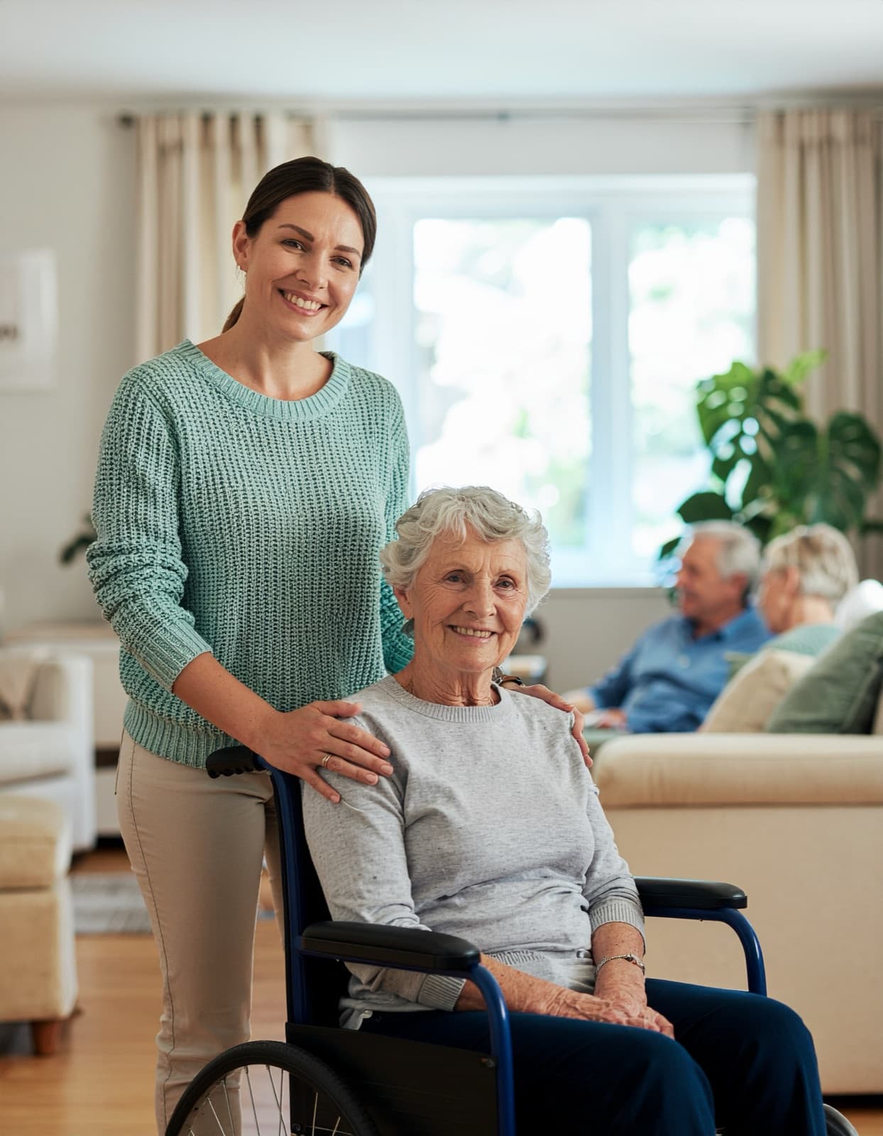Smiling caregiver standing beside an elderly woman in a wheelchair in the sunlit common living room of an adult care home