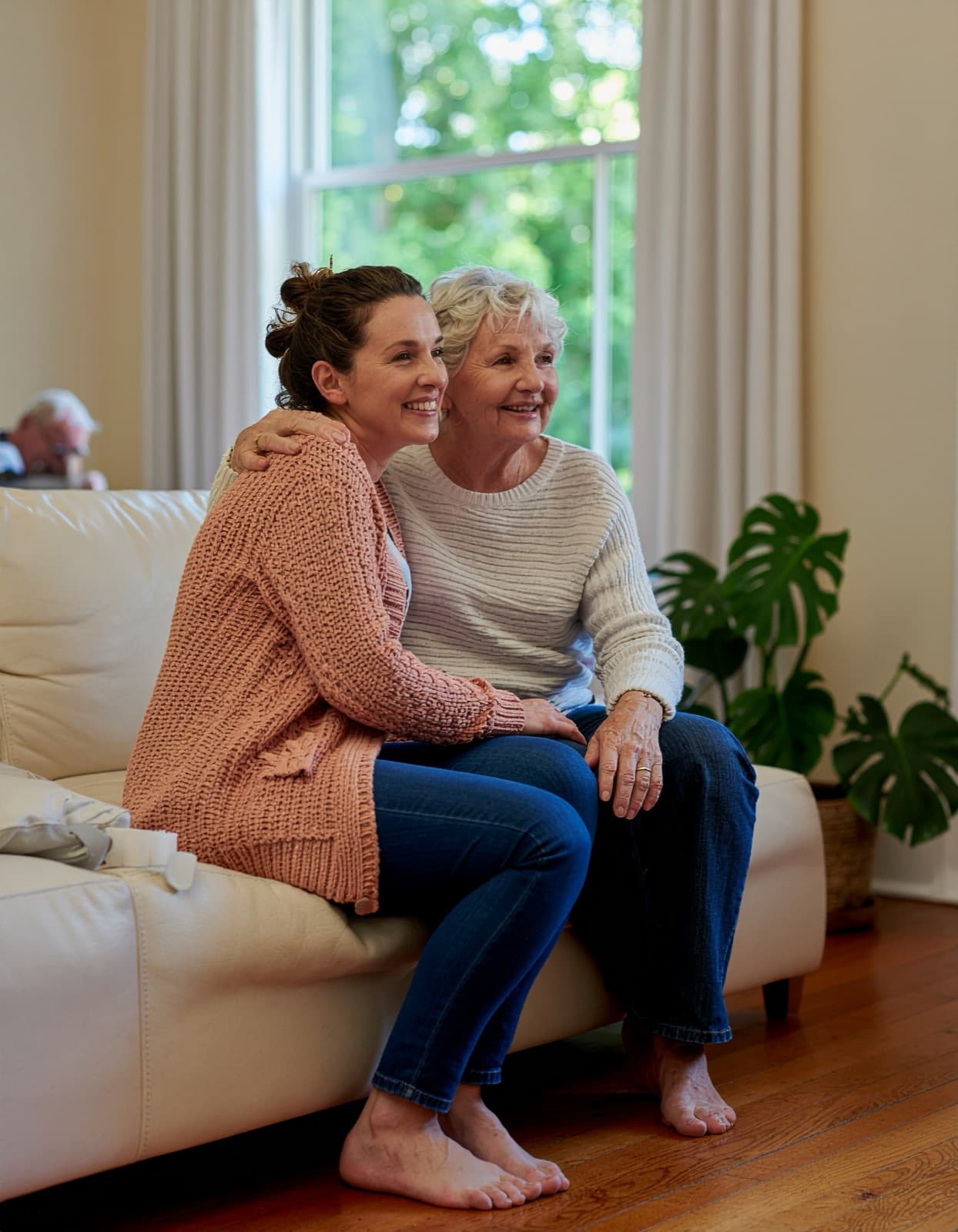 Adult daughter with her arm around her smiling elderly mother on a sofa in the sunlit common room of an adult care home