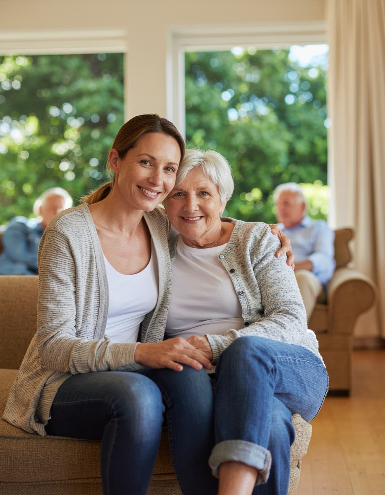Adult daughter sitting close beside her smiling elderly mother, holding hands, in the sunlit common room of an adult family home